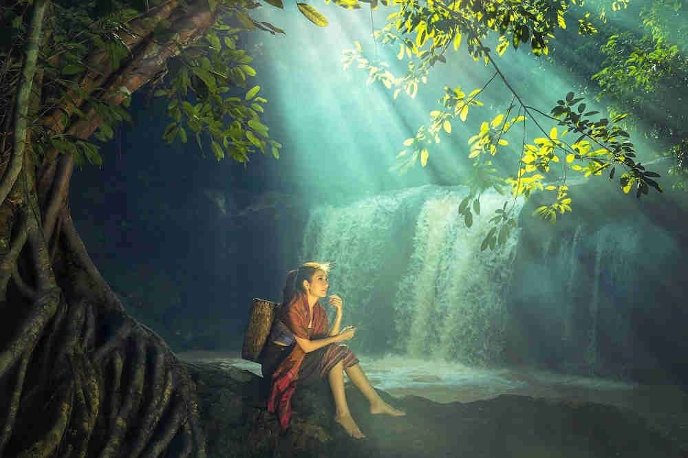 Woman sitting looking contemplative, below a sinewy-trunked tree with branches and leaves above her, near the edge of the water from a wide waterfall, brightened by diagonal sun-rays.